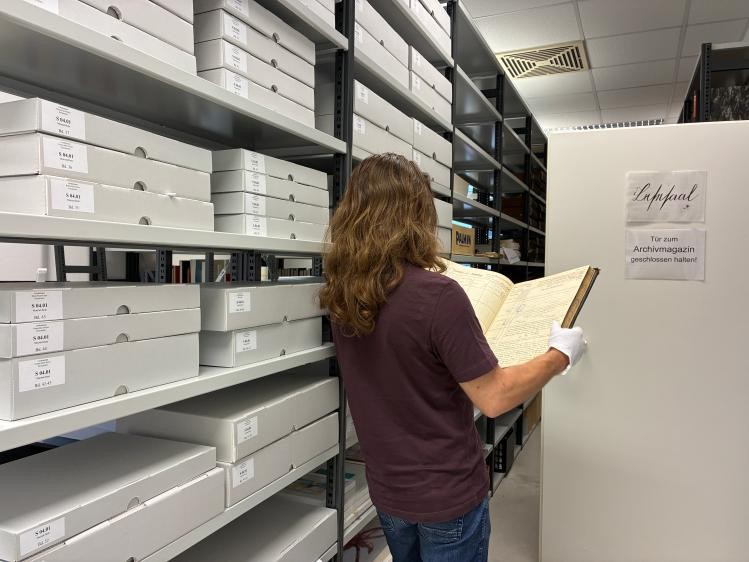 Eine Frau steht mit weißen Handschuhen und einem alten Buch in der Hand vor einem Archivregal
